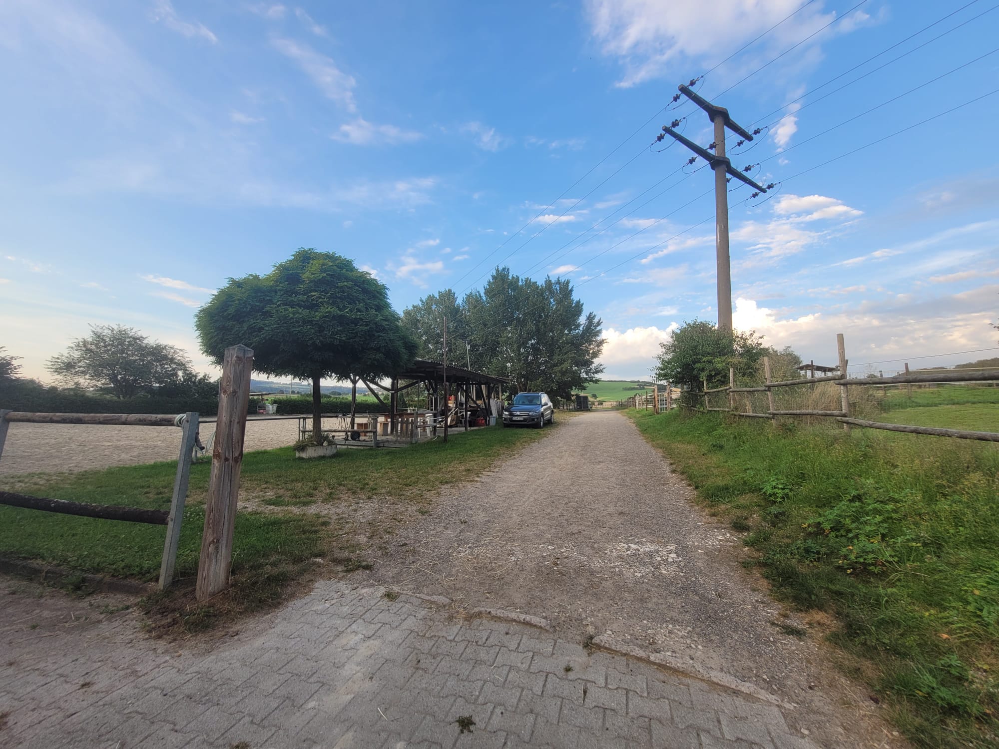 Ein Feldweg führt zu einer rustikalen Unterbringung und einem Auto, eingerahmt von Bäumen und offenen Feldern unter einem blauen Himmel mit Wolken, der die Ruhe dieser Startseite einfängt.