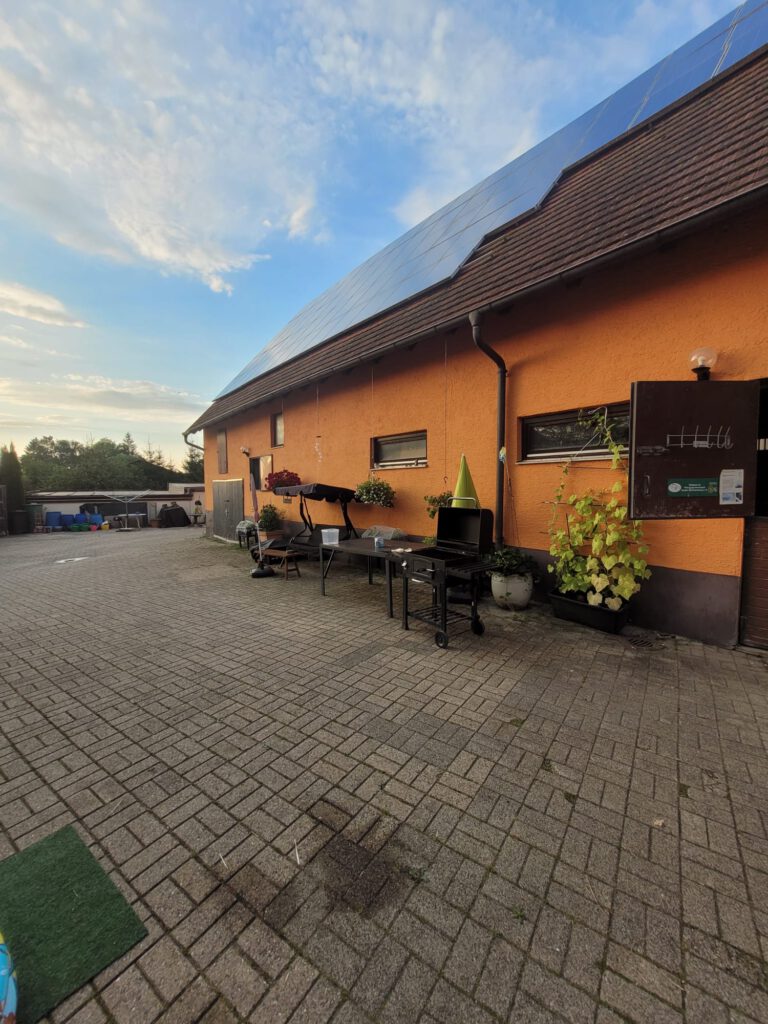 Ein gemauerter Innenhof mit Tisch, Grill und Pflanzen neben einem orangefarbenen Haus mit Sonnenkollektoren auf dem Dach heißt Sie auf Ihrer Startseite willkommen.