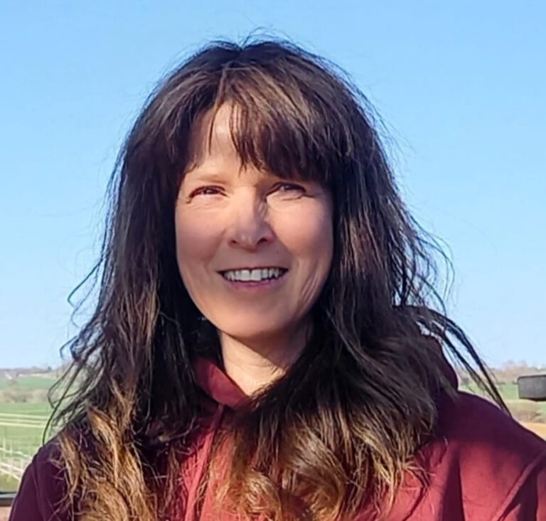 Eine Frau mit langem, welligem braunem Haar lächelt im Freien und trägt einen kastanienbraunen Kapuzenpullover. Blauer Himmel und eine verschwommene Landschaft im Hintergrund begrüßen Sie als perfekte Startseitenszene.