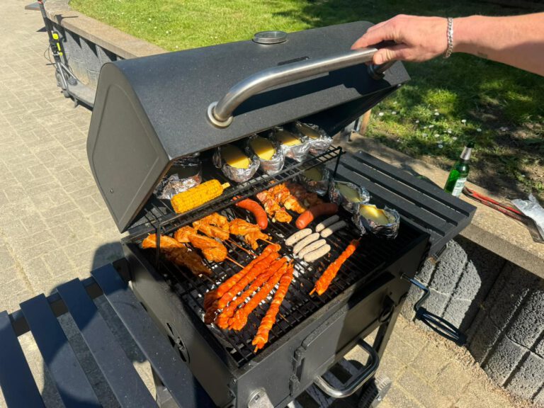 Person grillt verschiedene Fleischsorten, Mais und Kartoffeln auf einem großen schwarzen Holzkohlegrill vor einem Parkhintergrund und bereitet sich auf das Grillfest im Stall am 11. und 12. Mai 2024 vor.