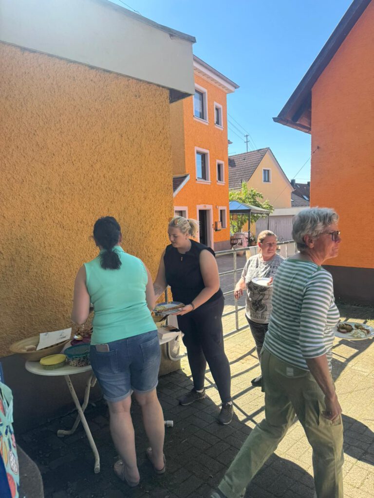 Menschen versammeln sich an einem sonnigen Tag draußen in einem Wohngebiet um einen Tisch und essen.