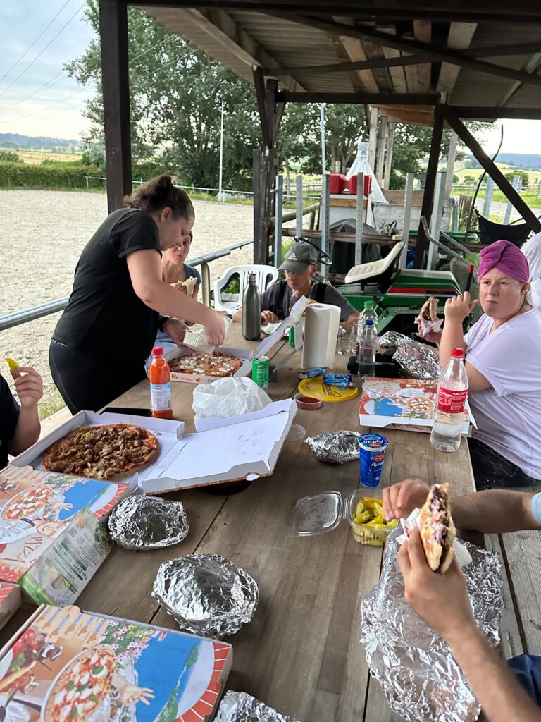 Eine Gruppe von Menschen genießt eine Mahlzeit im Freien. Speisen und Getränke sind auf einem Holztisch unter einer überdachten Konstruktion verteilt und feiern ihre erste Heuernte.