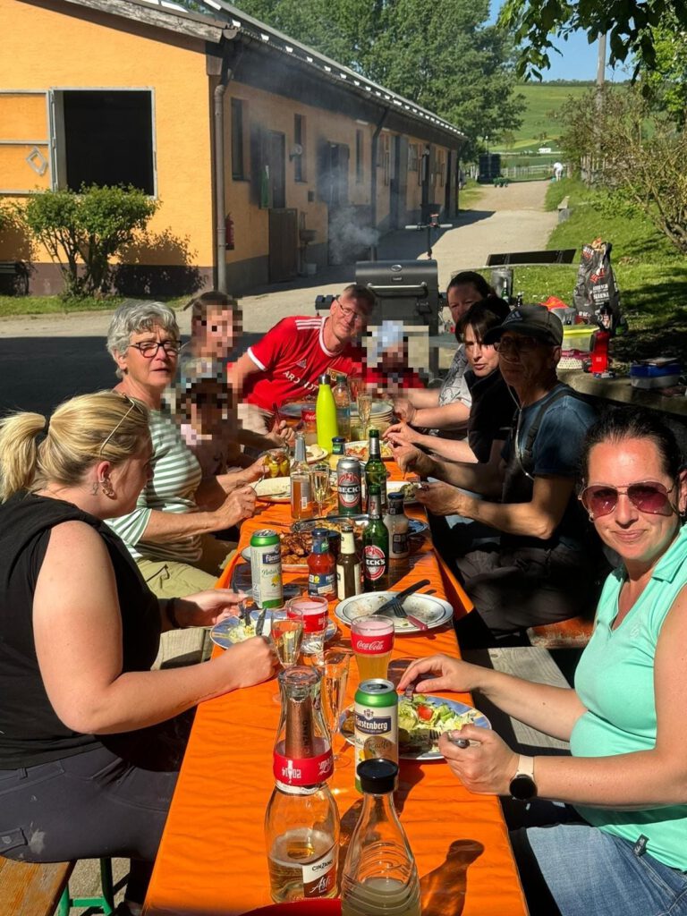 Eine Gruppe von Menschen genießt ein Essen im Freien an einem langen Tisch mit Getränken und Essen neben einem gelben Gebäude.