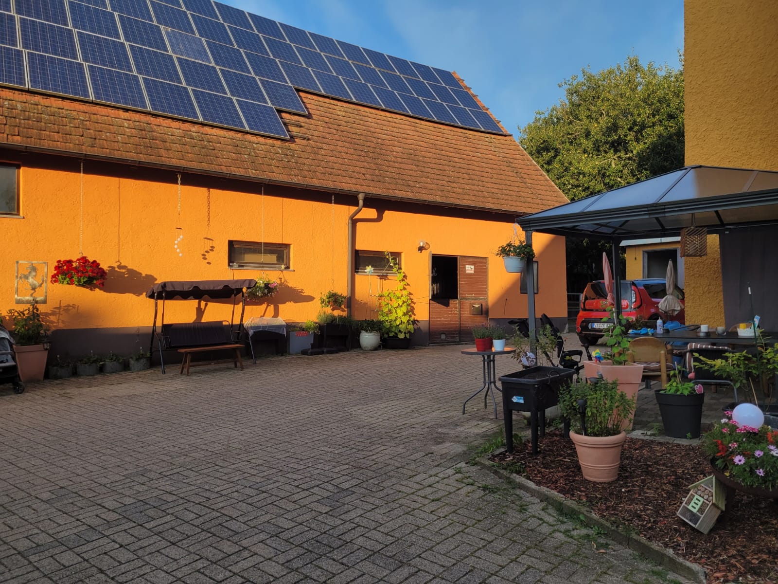 Eine Terrasse mit Topfpflanzen, einer Hollywoodschaukel und einem Haus mit Sonnenkollektoren auf dem Dach heißen Sie an einem sonnigen Tag in dieser ruhigen Startseite willkommen.