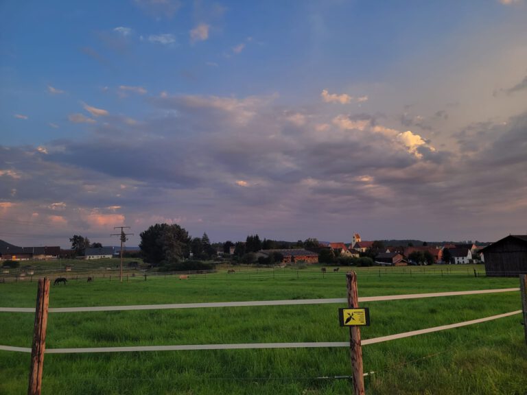 Eine ruhige ländliche Landschaft mit einer grünen Wiese, Holzzäunen, Häusern in der Ferne und einem Himmel mit vereinzelten Wolken bei Sonnenuntergang.