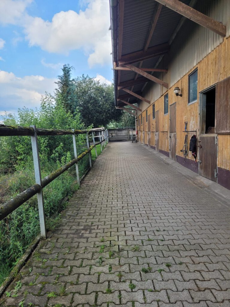 Ein Weg entlang eines Stalles mit Holztüren, Metallgeländern und Grünpflanzen unter einem teilweise bewölkten Himmel.