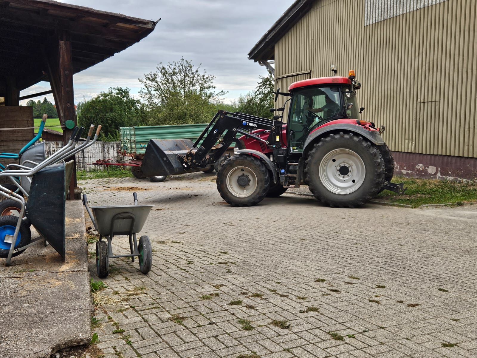 Ein roter Traktor mit Frontlader steht neben einer Schubkarre und einigen Geräten und dient als perfekte Startseite für alle landwirtschaftlichen Arbeiten außerhalb des Gebäudes.