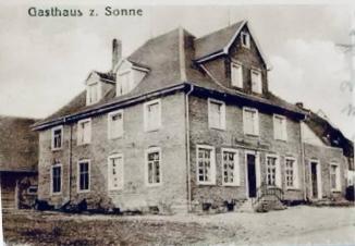 Sepiafarbenes Bild eines historischen Backsteingebäudes mit der Aufschrift „Gasthaus z. Sonne“ (Hotel zur Sonne) mit mehreren Fenstern und einem Satteldach, das die zeitlose Eleganz zeigt, die das Erbe von „über uns“ ausmacht.