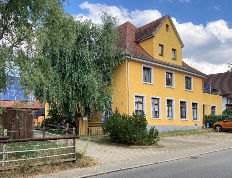 Ein charmantes, zweistöckiges gelbes Haus mit Satteldach, umgeben von Bäumen und einer kleinen Auffahrt, das in der Sonne liegt – erleben Sie die Wärme und den Komfort, die unseren charakteristischen Stil ausmachen. Über uns, es ist nicht nur ein Zuhause; hier werden Erinnerungen geschaffen.