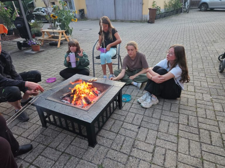 Vier Personen sitzen um eine Feuerstelle herum, eine Person hält eine Tasse und eine andere röstet Marshmallows, und erstellen so einen Entwurf gemütlicher Erinnerungen.