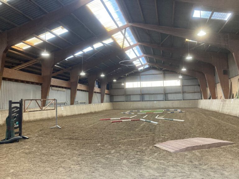 Indoor-Reithalle mit Hindernissen auf Sandboden, Holzbalken und natürlichem Licht durch Oberlichter.